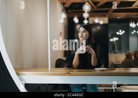Bella giovane donna che guarda via mentre si siede con la tazza di caffè pensando al caffè Foto Stock