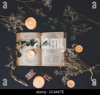 Una posa piatta di un libro vintage aperto su una superficie nera del tavolo con candele accese bianche e fiori secchi. Atmosfera intima e romantica Foto Stock