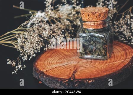 Primo piano di erbe di fireweed essiccate (willoweb) in un vaso quadrato di vetro. Bottiglia su piattaforma di legno tronco albero, con fiori di alito del bambino asciugato Foto Stock