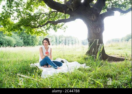 Donna giovane premurosa che guarda via mentre si siede con la mela su coperta da picnic da albero al parco Foto Stock