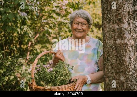 Sorridente donna anziana pensionato che tiene il cesto con fiori ed erbe mentre si levano in piedi dall'albero al giardino Foto Stock