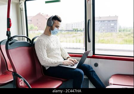 Giovane uomo che indossa una maschera protettiva seduto in autobus pubblico con cuffie e computer portatile, Spagna Foto Stock