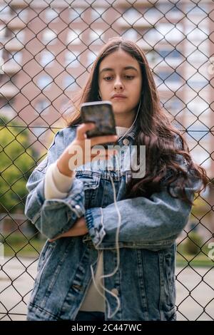 Ragazza adolescente sicura che usa lo smartphone contro la recinzione chainlink al campo sportivo Foto Stock