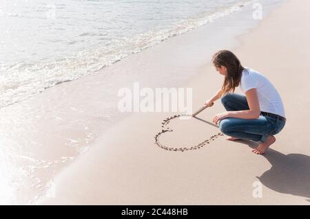 Donna accovacciata sulla spiaggia grattando cuore in sabbia bagnata con bastone di legno, Sardegna, Italia Foto Stock