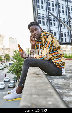 Giovane uomo in camicia a motivi posti sulla terrazza sul tetto della città che parla al telefono, Maputo, Mozambico Foto Stock