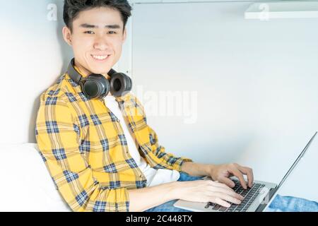 Ritratto di un giovane sorridente con le cuffie seduti a letto utilizzando un computer portatile Foto Stock