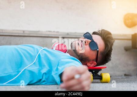 Ritratto di uomo che indossa occhiali da sole sdraiato a terra con la testa sullo skateboard Foto Stock