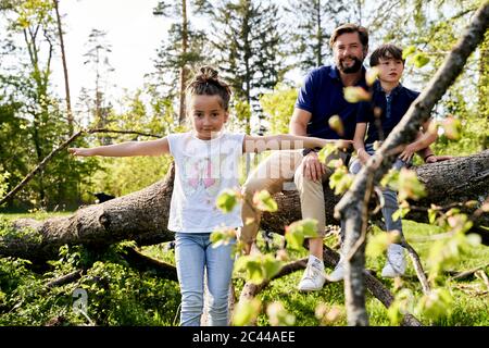 Ragazza carina con le braccia allungate in piedi contro padre e fratello seduto su albero caduto in foresta Foto Stock