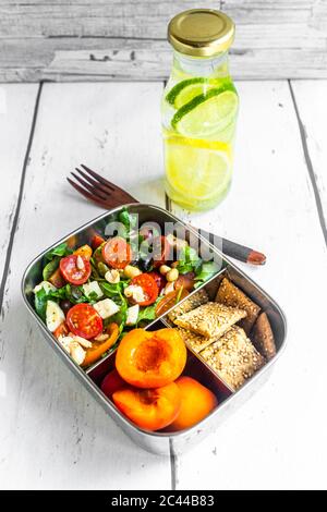 Pranzo al sacco con insalata di razzi con pomodori colorati, mozzarella e noci, croccante e albicocche e bottiglia d'acqua infusa con limoni e fette di lime Foto Stock