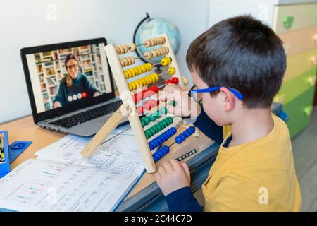 Studente maschile con abaco alla scrivania mentre insegnante in videochiamata durante la COVID-19 homeschooling Foto Stock