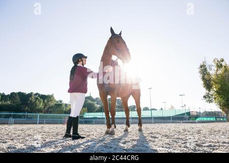 Ragazza teenage illuminata con cavallo di castagno su terreno di allenamento contro il cielo limpido Foto Stock