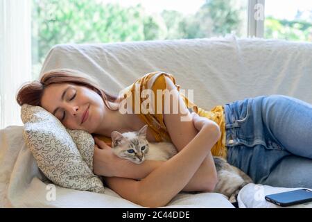 Giovane donna con gli occhi chiusi che si stendeva sul divano a casa con il suo gatto Foto Stock