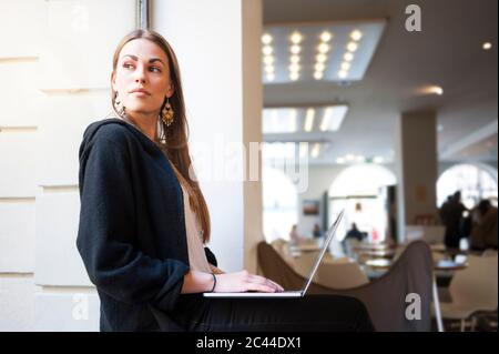 Donna premurosa che guarda attraverso la finestra mentre usa il computer portatile nella caffetteria Foto Stock