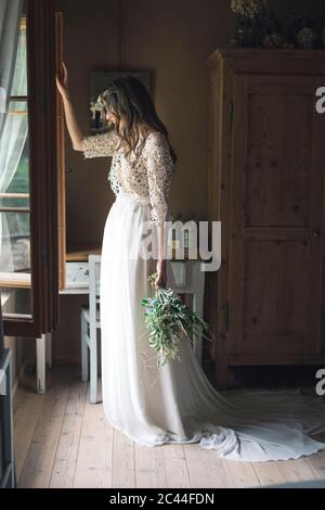 Giovane donna in elegante abito da sposa che tiene bouquet che guarda fuori dalla finestra Foto Stock