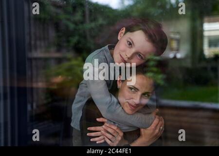 Figlio amorevole abbracciando la madre mentre si sta a casa visto attraverso la finestra Foto Stock