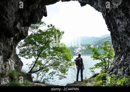 Uomo escursionista guardando il Lago di Como contro il cielo dall'ingresso della grotta Foto Stock