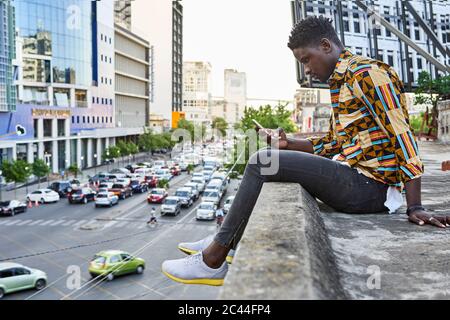 Giovane uomo in camicia a motivi posti sulla terrazza sul tetto della città controllare il suo telefono, Maputo, Mozambico Foto Stock