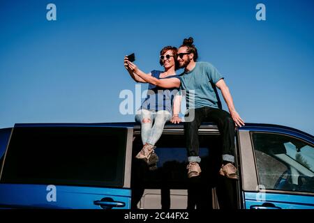 Felice coppia seduta sul tetto di un minivan prendendo un selfie Foto Stock