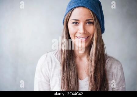 Donna felice con lunghi capelli marroni che indossa cappello a maglia contro parete grigia Foto Stock