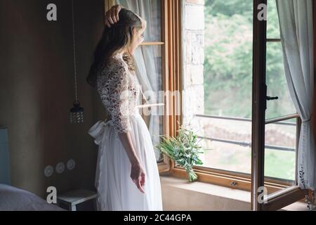 Giovane donna in elegante abito da sposa che tiene bouquet che guarda fuori dalla finestra Foto Stock