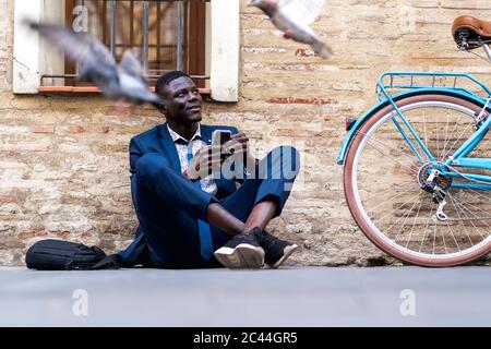 Giovane uomo d'affari con smartphone seduto a terra in città Foto Stock