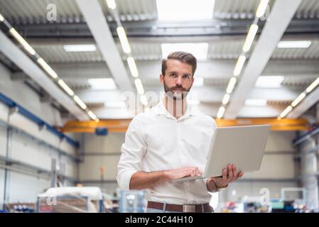 Giovane uomo che tiene il computer portatile sul pavimento della fabbrica Foto Stock