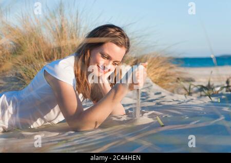 Ritratto di donna sorridente sdraiata sulla spiaggia che le insidiava la sabbia con la mano, Sardegna, Italia Foto Stock