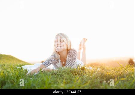 Felice giovane donna pensierosa che si stese su terra erbosa contro il cielo limpido nel parco durante il tramonto Foto Stock