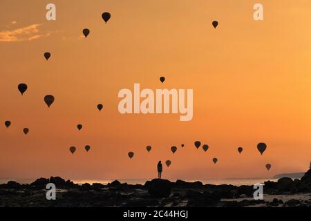 Indonesia, Nusa Tenggara occidentale, Silhouette di mongolfiere che volano sopra la donna solita in piedi sulla costa rocciosa al tramonto della moody Foto Stock