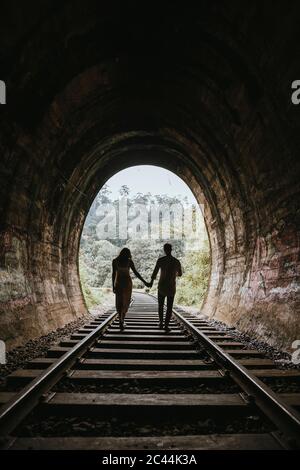 Sri Lanka, Provincia di Uva, Demodara, Silhouette di coppia che tiene le mani mentre si cammina giù tunnel che conduce al Ponte Nine Arch Foto Stock