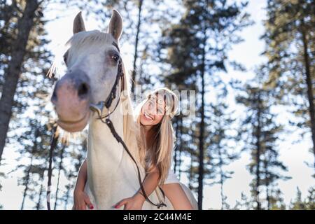 Vista ad angolo basso di una giovane donna sorridente appoggiata a cavallo bianco nella foresta Foto Stock