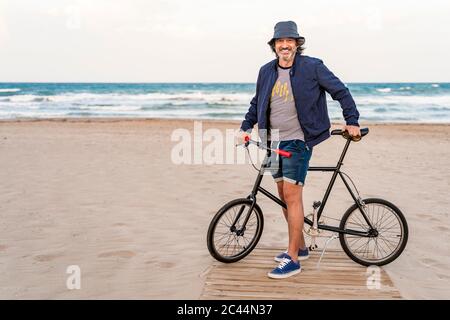 Uomo maturo con bicicletta, in piedi sulla spiaggia, sorridente Foto Stock