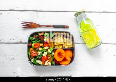 Pranzo al sacco con insalata di razzi con pomodori colorati, mozzarella e noci, croccante e albicocche e bottiglia d'acqua infusa con limoni e fette di lime Foto Stock