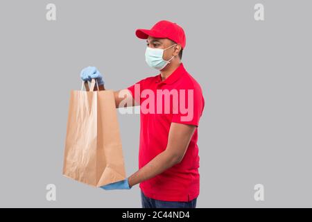 Uomo di consegna con sacchetto di carta in mani che indossa maschera medica e guanti guardando lato isolato. Ragazzo di consegna indiano uniforme rosso. Consegna di cibo domestico Foto Stock