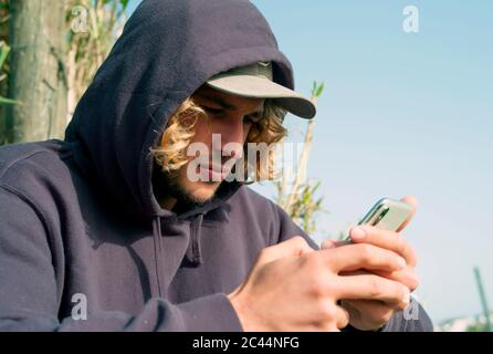 Primo piano di un giovane uomo che indossa il cappuccio utilizzando uno smartphone mentre si rilassa alla spiaggia Tarifa, Spagna Foto Stock