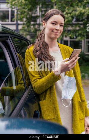 Ritratto di giovane donna sorridente con maschera protettiva e cellulare appoggiato in auto Foto Stock