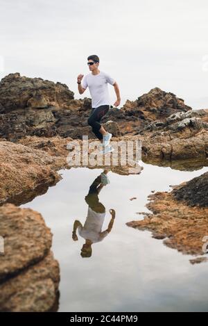 Lunghezza completa di corridore di sentiero maschile che corre su rocce con il suo riflesso sull'acqua a costa, Ferrol, Galizia, Spagna Foto Stock