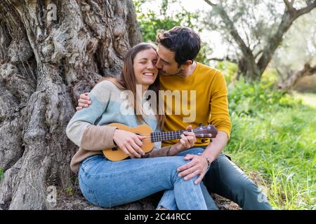 Ragazzo romantico baciare felice donna che suona la chitarra mentre si siede contro tronco d'albero Foto Stock