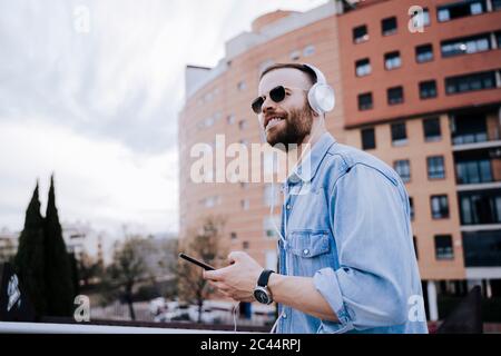 Ritratto di un giovane sorridente che ascolta musica con cuffie e smartphone all'aperto Foto Stock