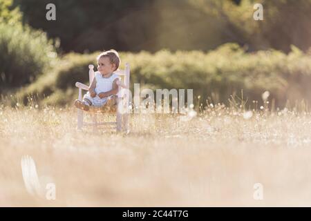 Carina bambina che guarda via mentre si siede su sedia sul prato durante la giornata di sole Foto Stock