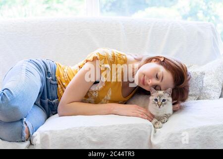 Giovane donna con gli occhi chiusi che si stendeva sul divano a casa con il suo gatto Foto Stock