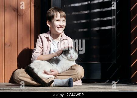 Ragazzo sorridente che guarda via mentre accarezzava gatto rilassandosi su di lui in portico durante il giorno di sole, Tarusa, Russia Foto Stock