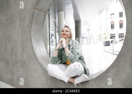 A piena lunghezza Ritratto di bella donna matura sorridente mentre è seduto sul cerchio di calcestruzzo nella parete Foto Stock