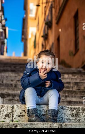 Una ragazza felice (caucasica) sorride e gode di sedersi su un gradino di una scala, indossando un cappotto blu, in una giornata di sole inverno. Scorcio del cielo blu b Foto Stock