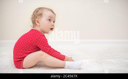 Bambina in una T-shirt rossa seduta su un tappeto bianco nella stanza. Foto Stock