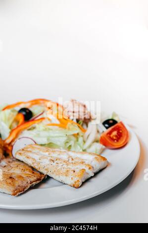 Bistecca di pesce persico dorato alla griglia con pomodoro fresco, lattuga e olive su piatto bianco e fondo bianco Foto Stock
