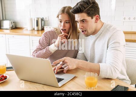 Ritratto di una giovane coppia scioccata che punta il dito sul computer portatile mentre fa colazione in una cucina accogliente a casa Foto Stock