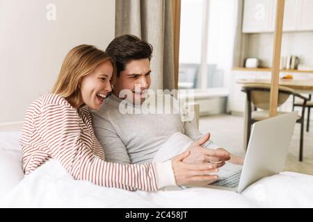 Ritratto di una bella coppia emozionata sorridente e che usa il computer portatile mentre si trova a letto a casa Foto Stock