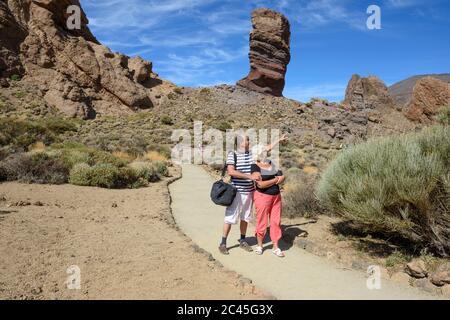 Coppie anziane si posano sul sentiero vicino Garcia Rocks con roccia di Cinchado sullo sfondo nel Parco Nazionale di Teide, Tenerife, Isole Canarie, Spagna. Foto Stock