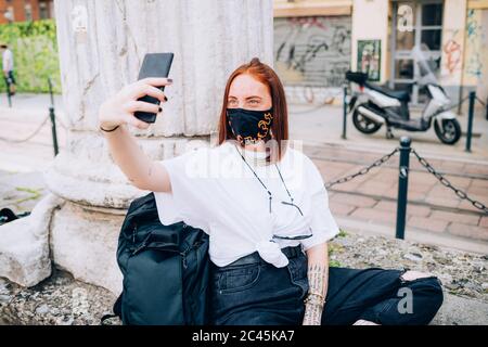 Giovane donna che indossa la maschera durante il virus Corona, seduta all'aperto, prendendo selfie. Foto Stock